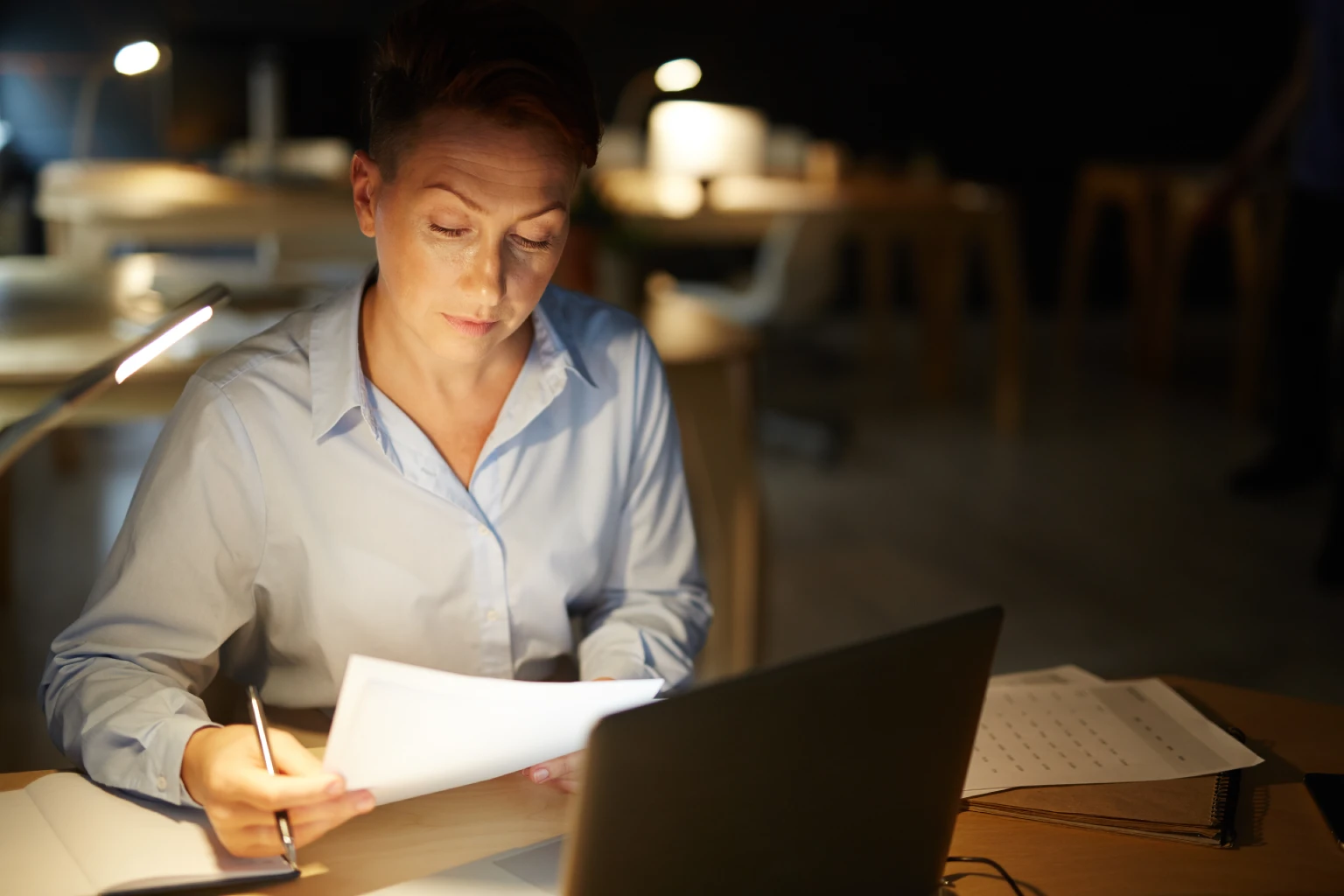 Mulher analisando documentos em uma mesa iluminada por luminária, à noite