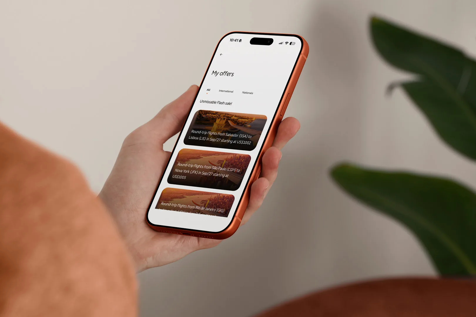 Mão segurando o celular com a tela de ofertas do app da GOL em inglês: voos de ida e volta para Lisboa e Nova York com preços em dólar